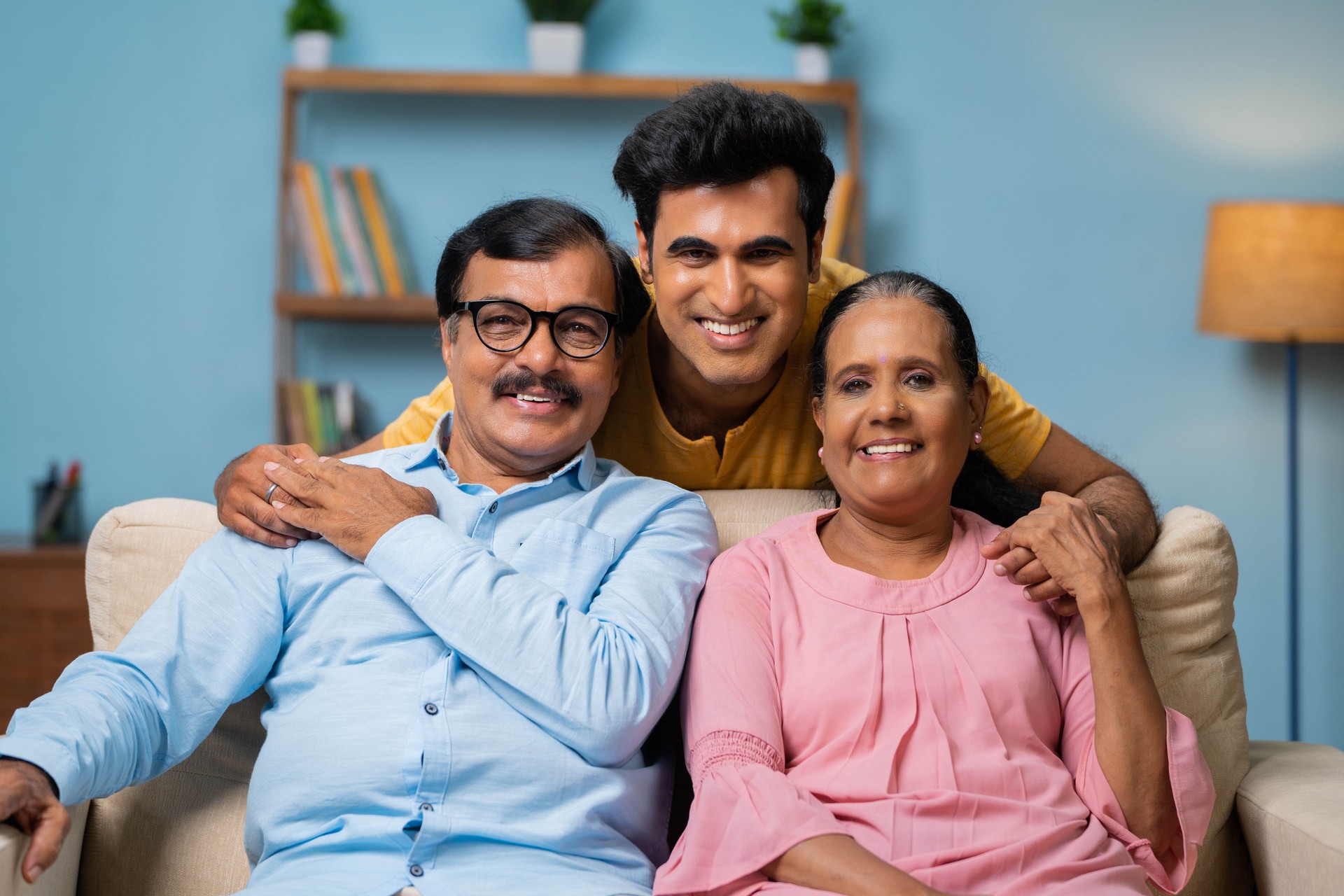 portrait shot of Happy smiling indian senior parents with adult son looking at camera while sitting on sofa at home - concept of family bonding, relationship and relaxation.